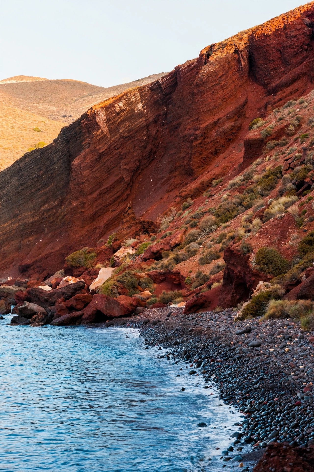 Dramatic rust-red volcanic cliffs rising behind the dark pebble shore of Red Beach near Akrotiri, Santorini