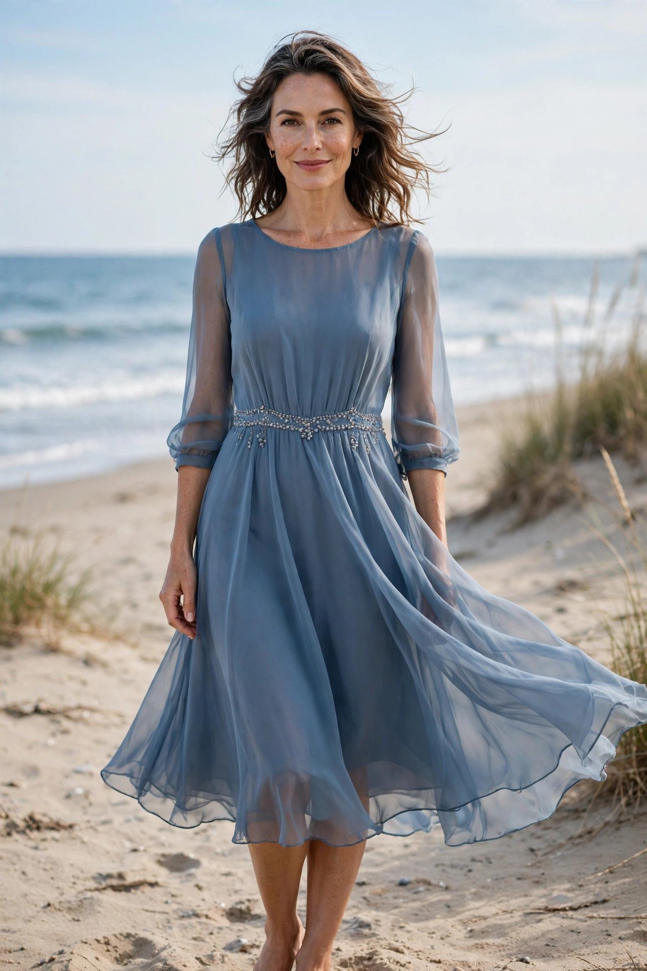 Beautiful woman in her late 40s in a dusty blue chiffon A-line dress with sheer sleeves on the beach