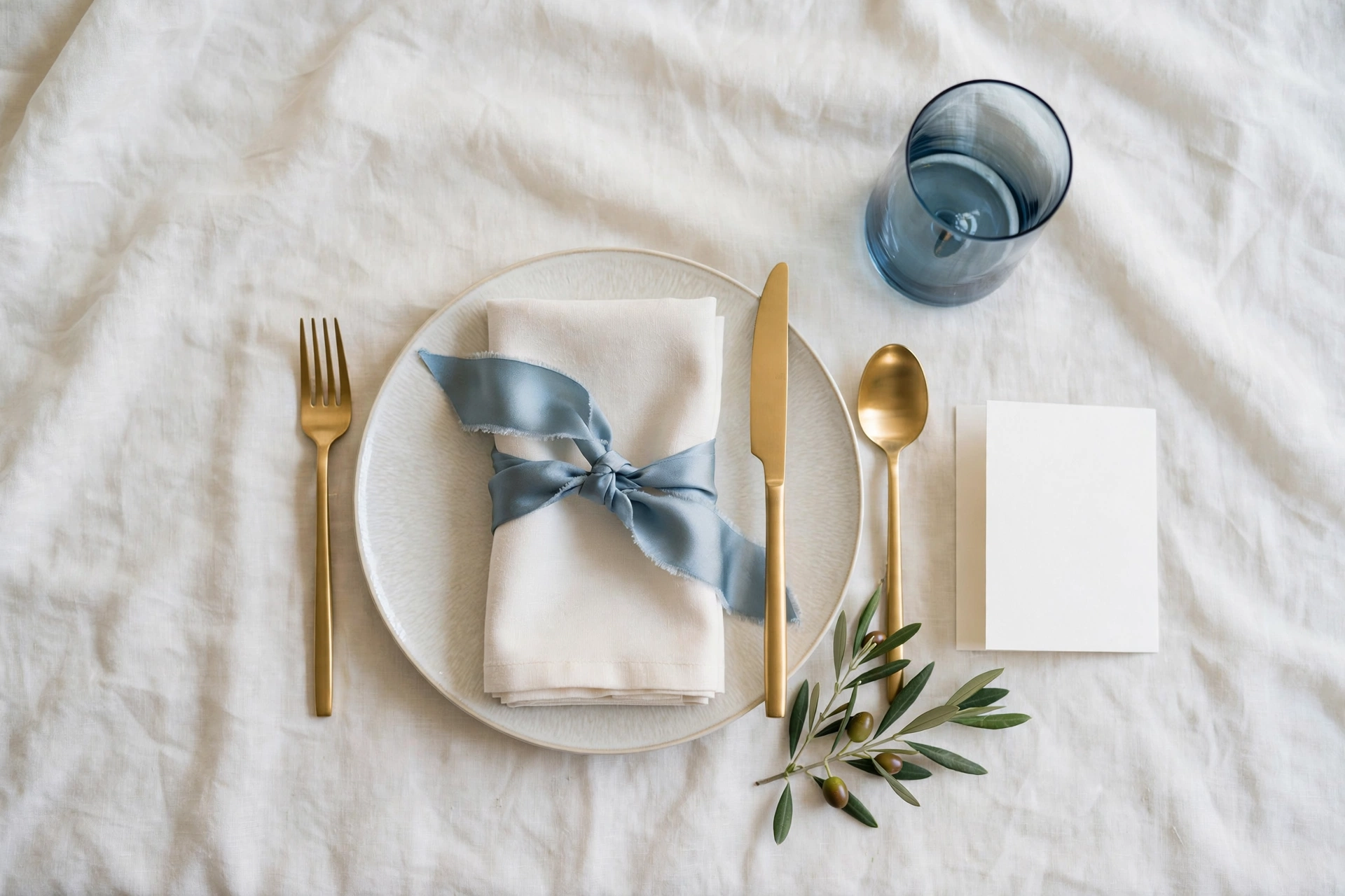 Elegant wedding place setting flat-lay with calligraphed menu card, gold flatware and dusty-blue glassware on ivory linen